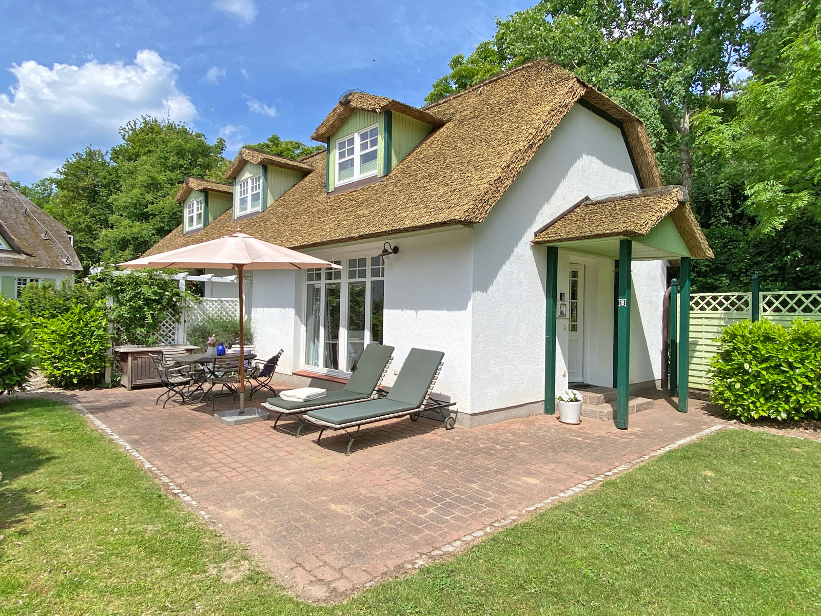 White cottage with a thatched roof, patio, lounge chairs, dining set, and umbrella in a lush green garden. [AI-generated text]