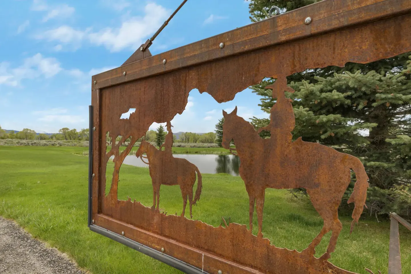 A rusted metal sign featuring cowboy silhouettes overlooks a green pasture, pond, and pine trees under a blue sky.