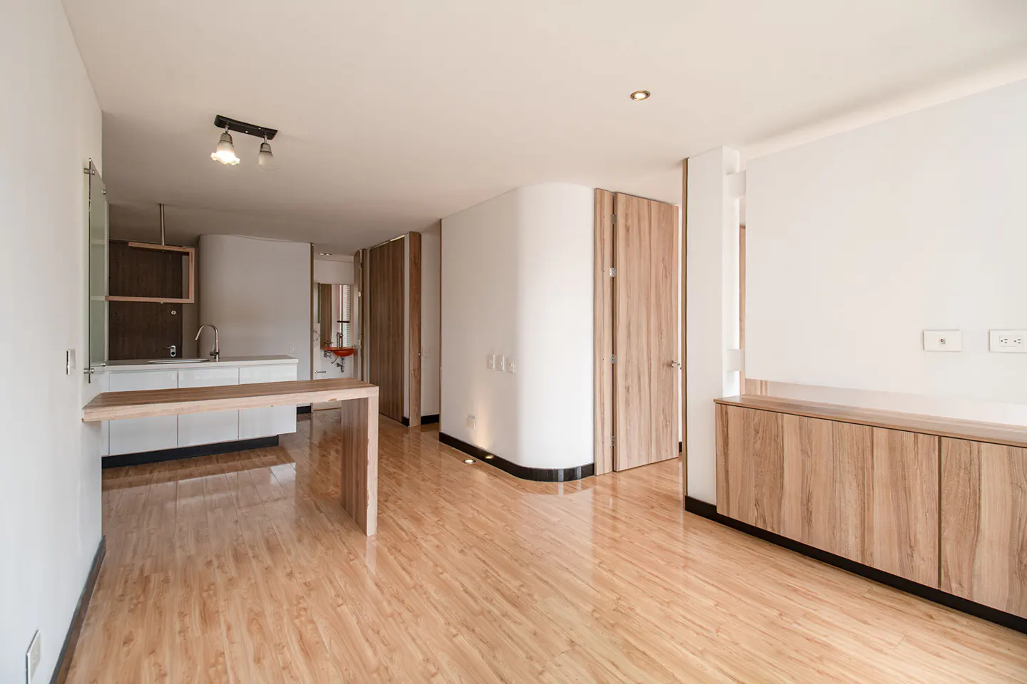 Bright, modern apartment interior with wood floors, white walls, and wood cabinets. A kitchen area is visible with a breakfast bar.