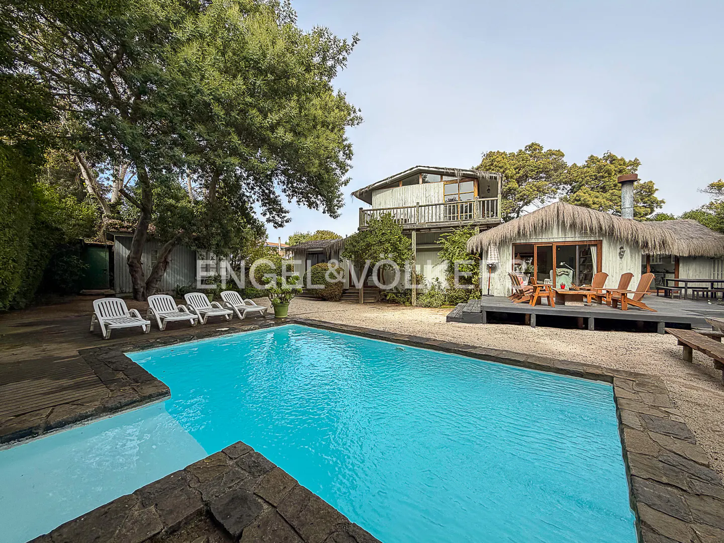 A bright blue swimming pool sits in front of a rustic house with a thatched roof, wooden deck, and lounge chairs.