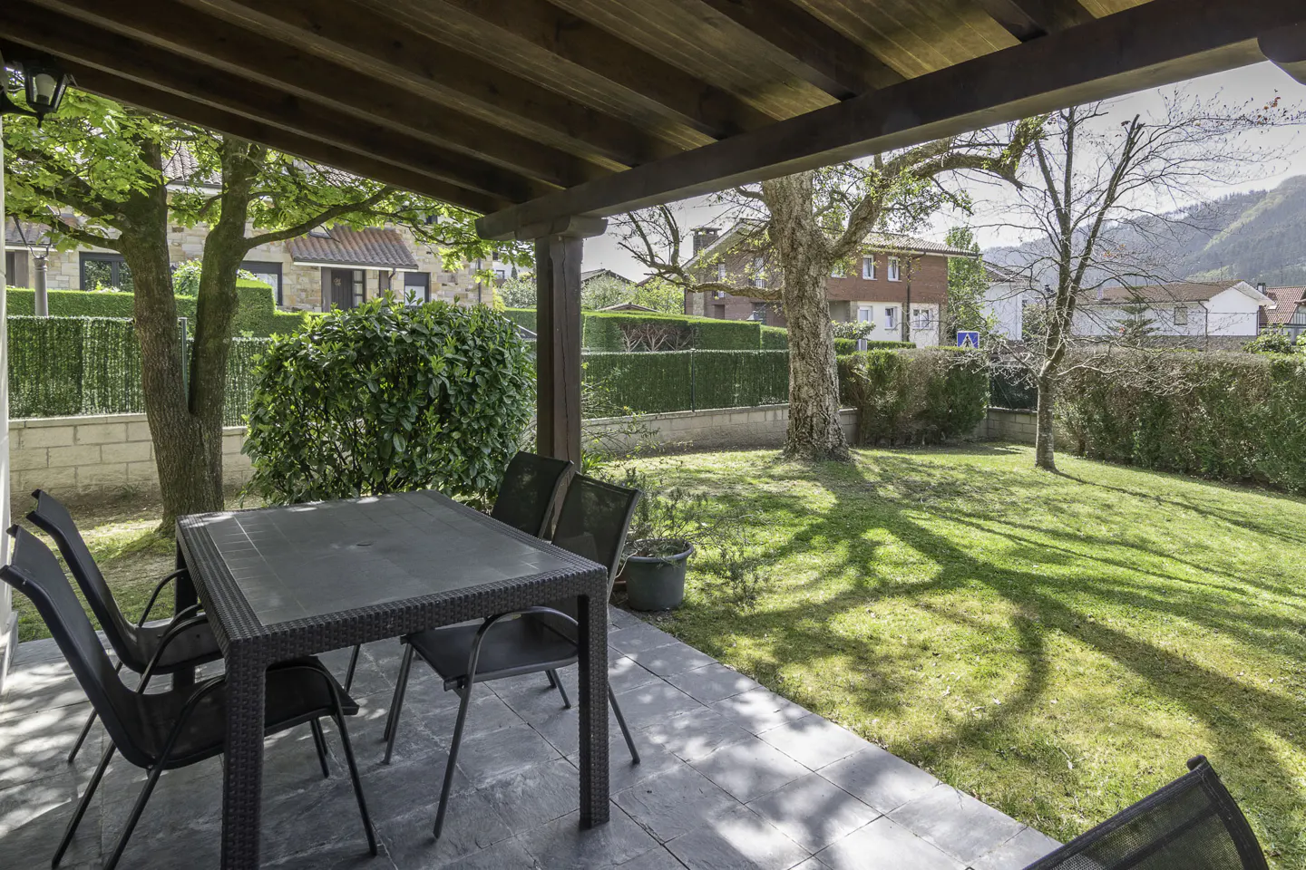 Covered patio with a black table and chairs overlooking a green lawn and trees in a residential area.