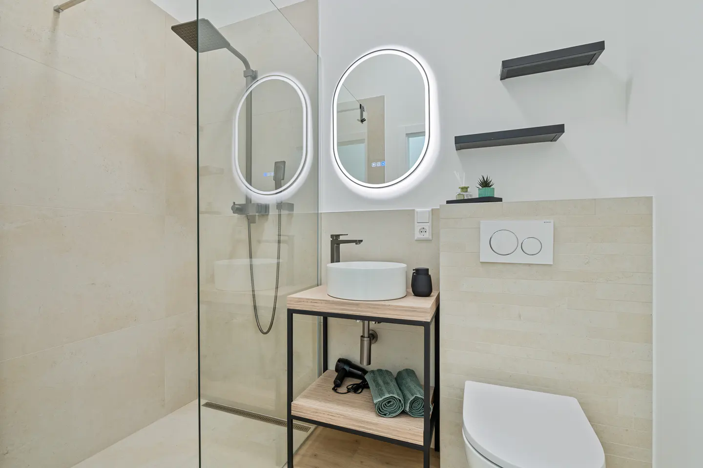 Modern bathroom with a glass shower, oval LED mirrors, a white sink on a wooden stand, and a toilet. Two black shelves are on the wall.
