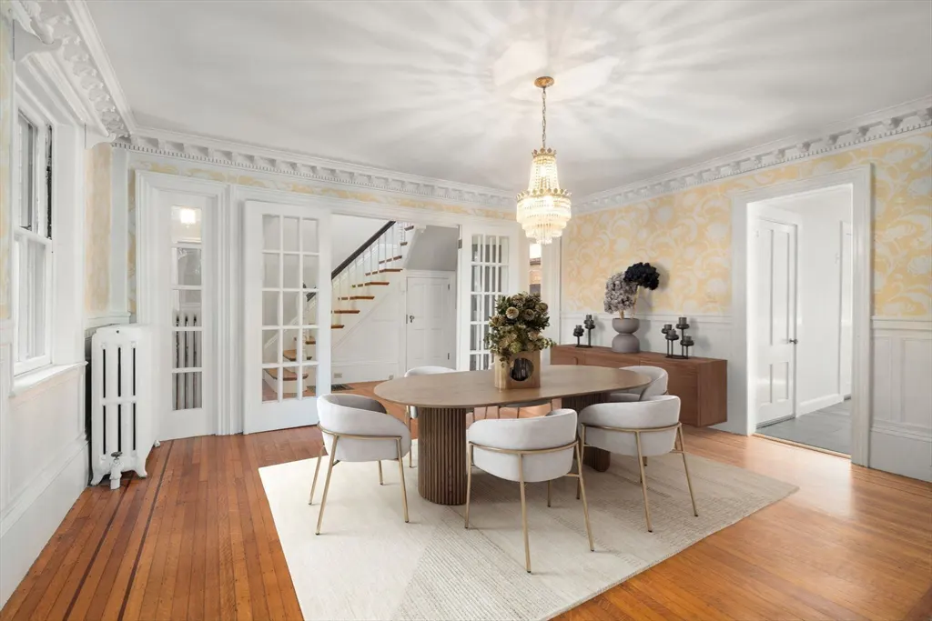 A dining room with hardwood floors, an oval table with six chairs, and a crystal chandelier. French doors lead to a staircase.