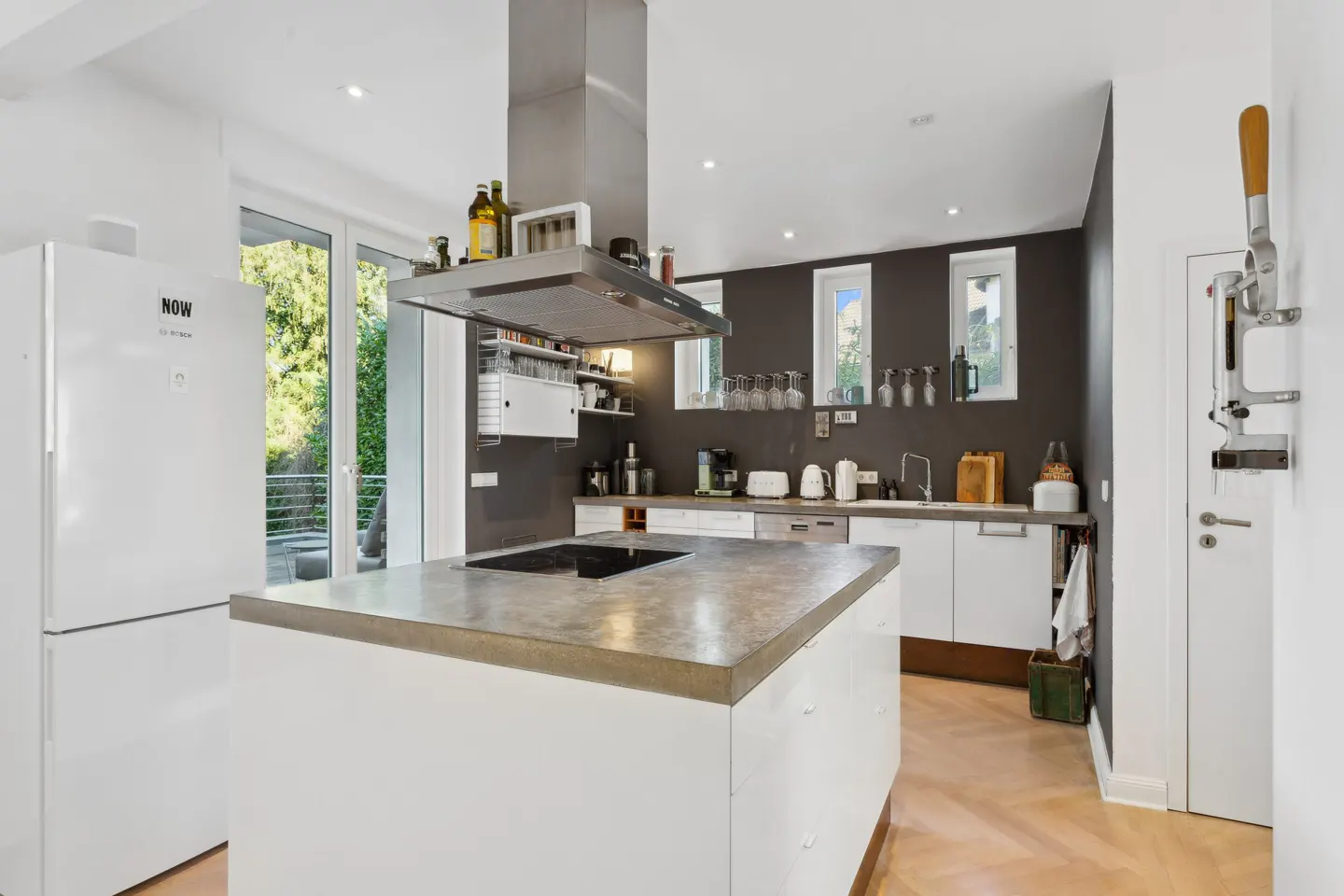 Bright kitchen with a white island, stainless steel hood, and white cabinets against a dark gray wall. A white refrigerator stands to the left.