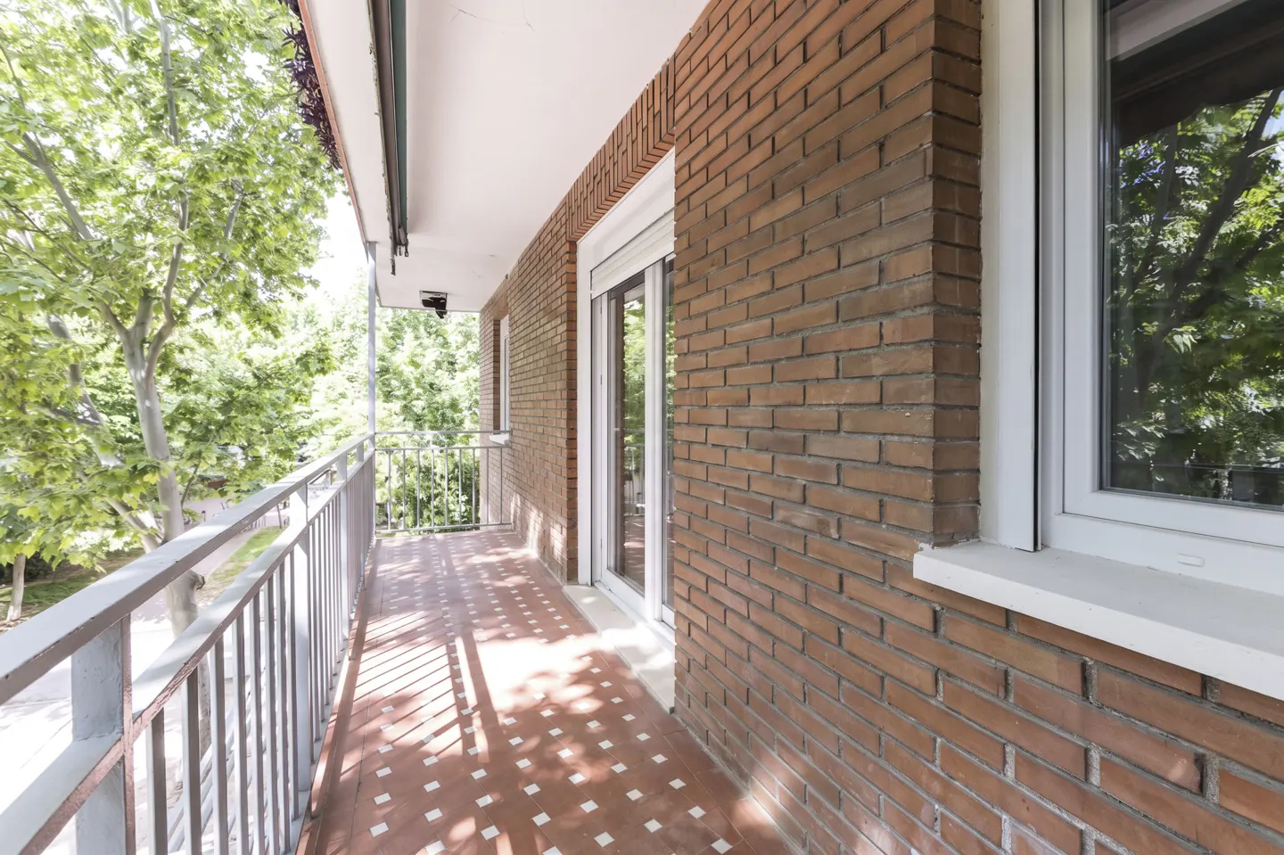 A long balcony with a gray metal railing, red tiled floor, and a brick wall. Green trees are visible outside.