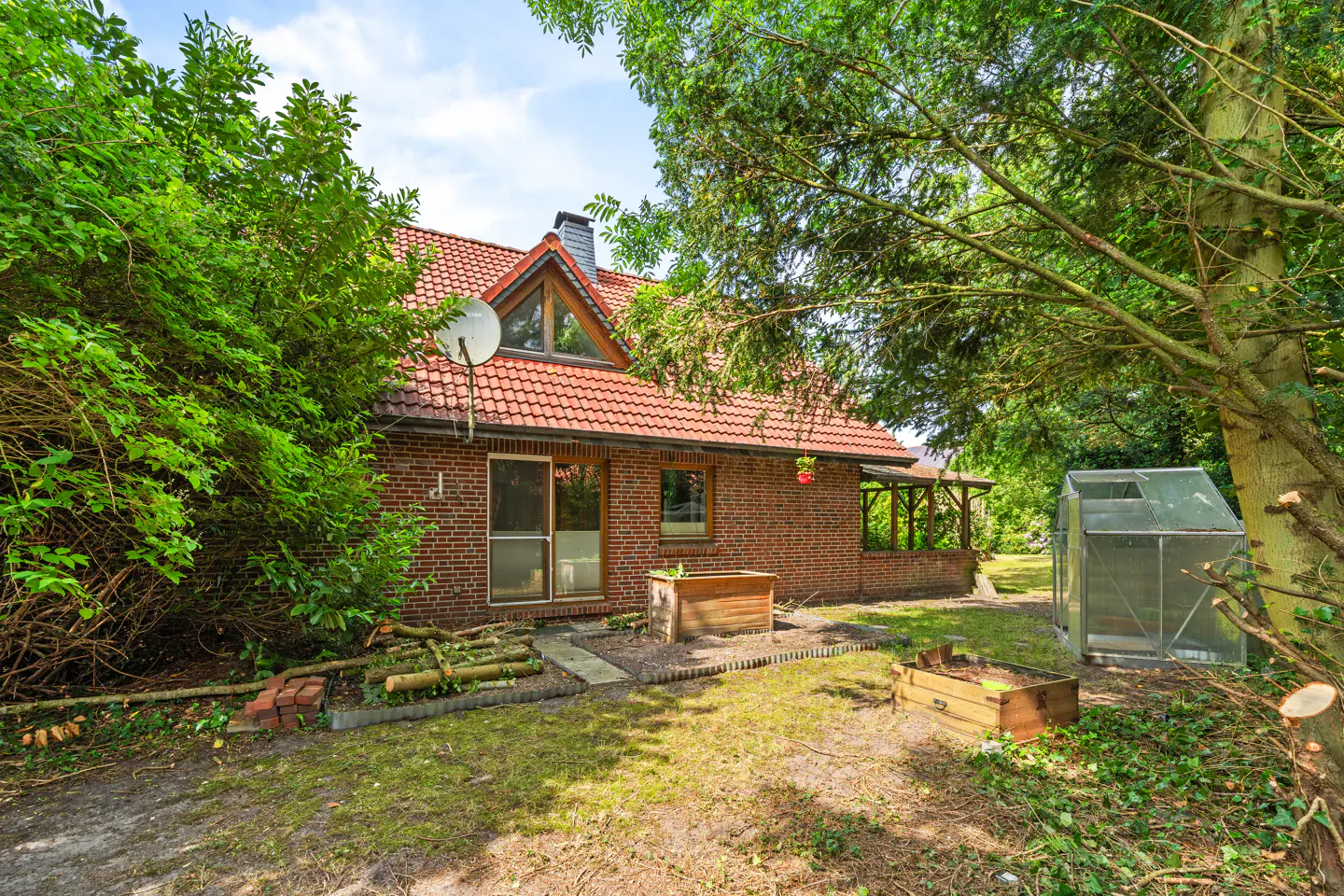 Red brick house with a tiled roof, surrounded by lush trees, a small greenhouse, and wooden garden planters in the yard.