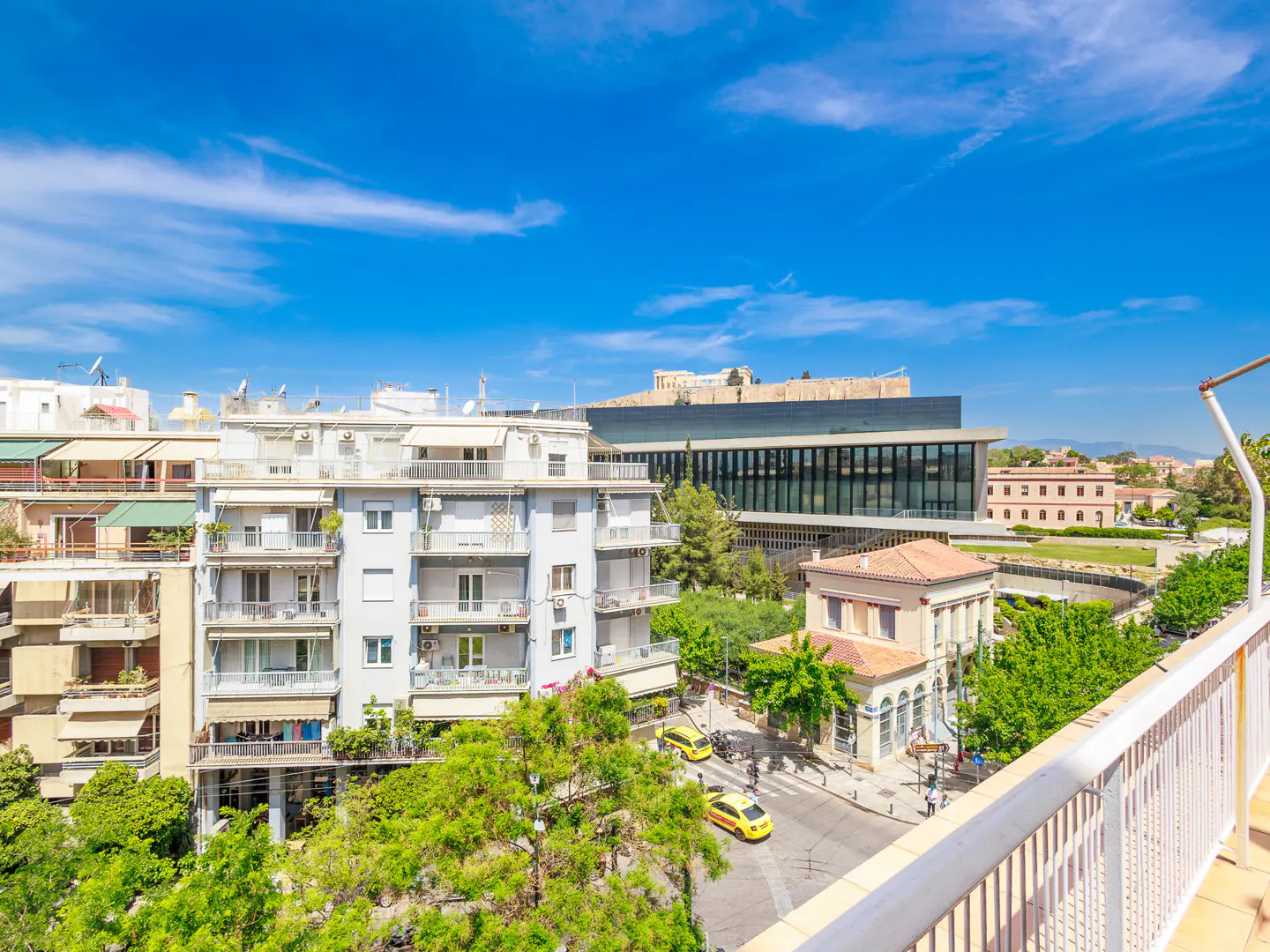 View from a balcony overlooking Athens, Greece, with buildings, trees, and the Acropolis in the distance under a blue sky.