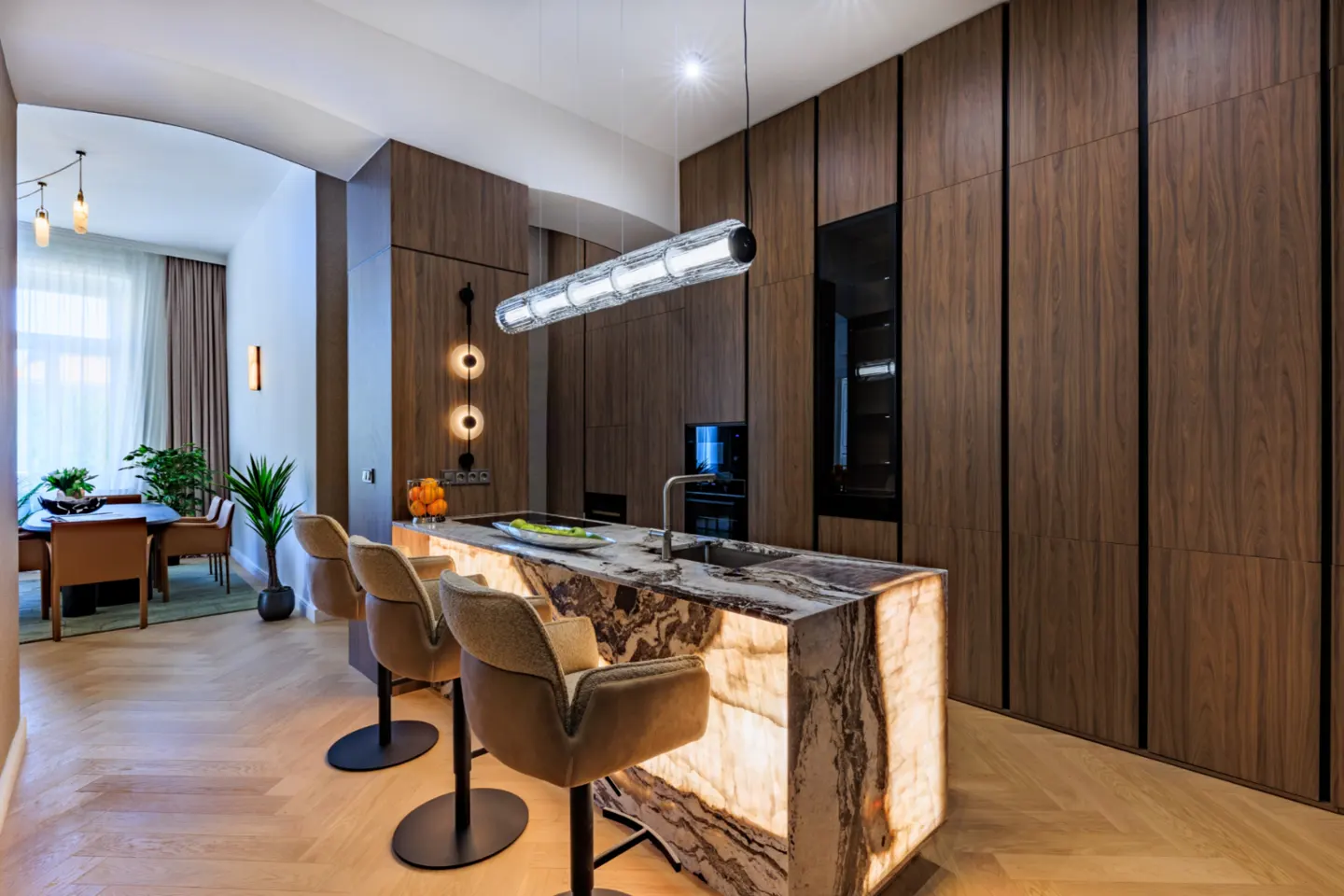 A modern kitchen with a marble island, brown cabinets, and three bar stools. A dining table is visible through an archway.