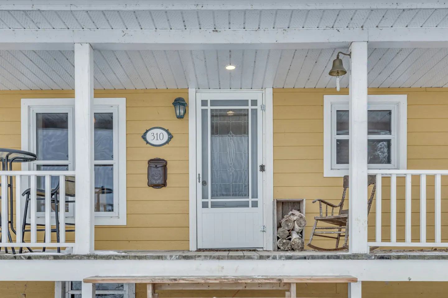 Front view of a yellow house with a white porch, door, and windows. A rocking chair and firewood are on the porch.