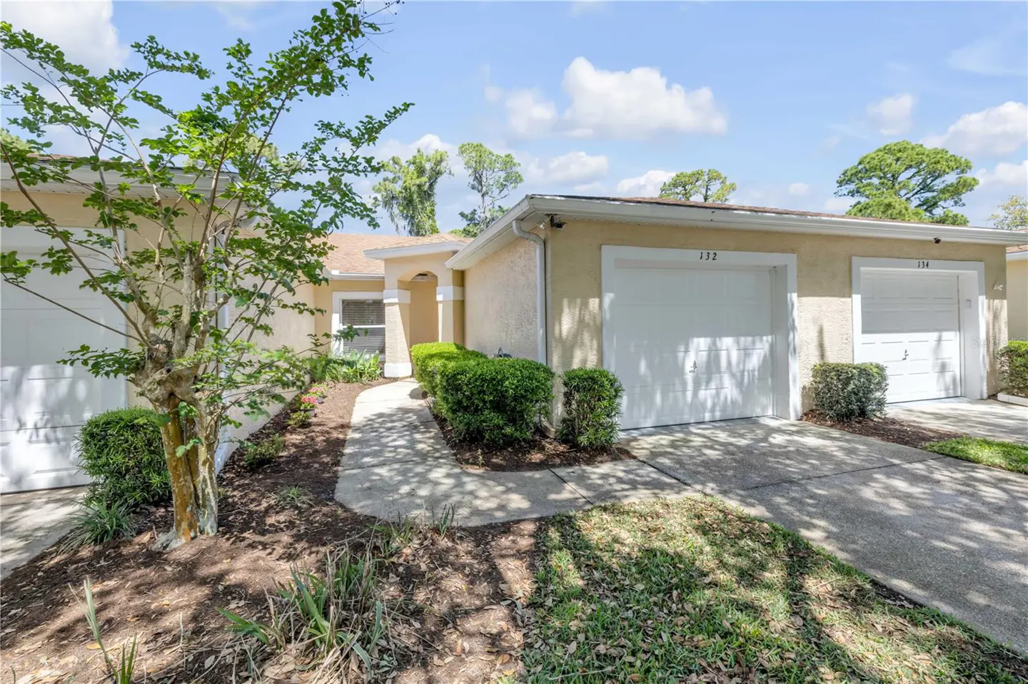 Exterior view of a one-story, beige house with white garage doors, green bushes, and a tree.