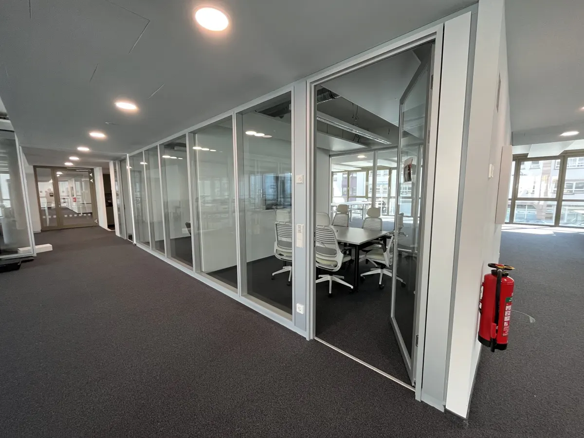 Office hallway with glass-walled meeting rooms. A conference table and chairs are visible inside. A red fire extinguisher is mounted on the wall.