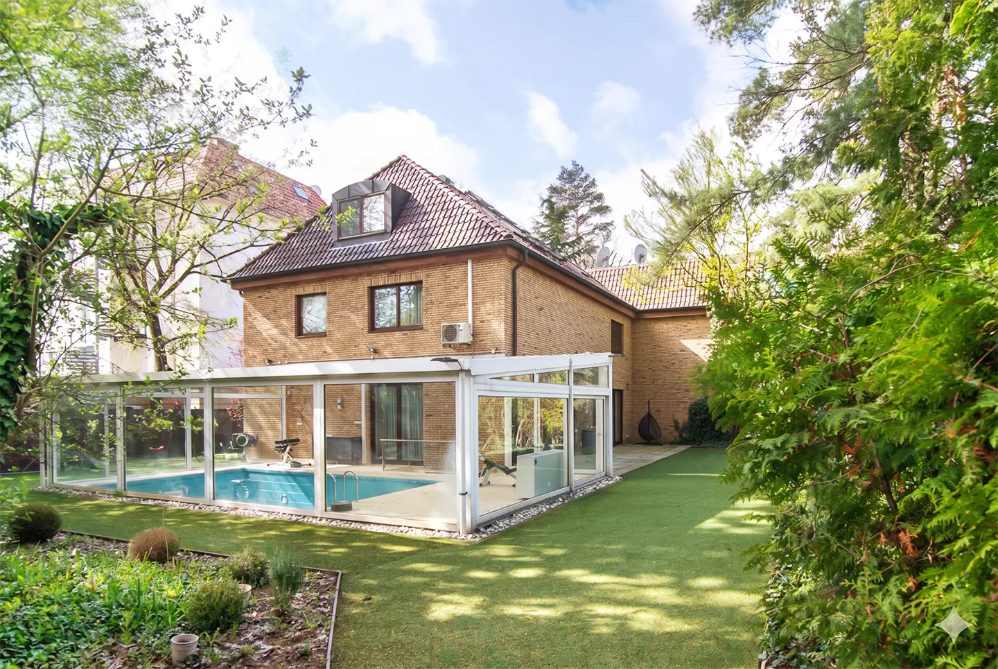 Brick house with a glass-enclosed pool and gym equipment, surrounded by green lawn and trees under a blue sky.