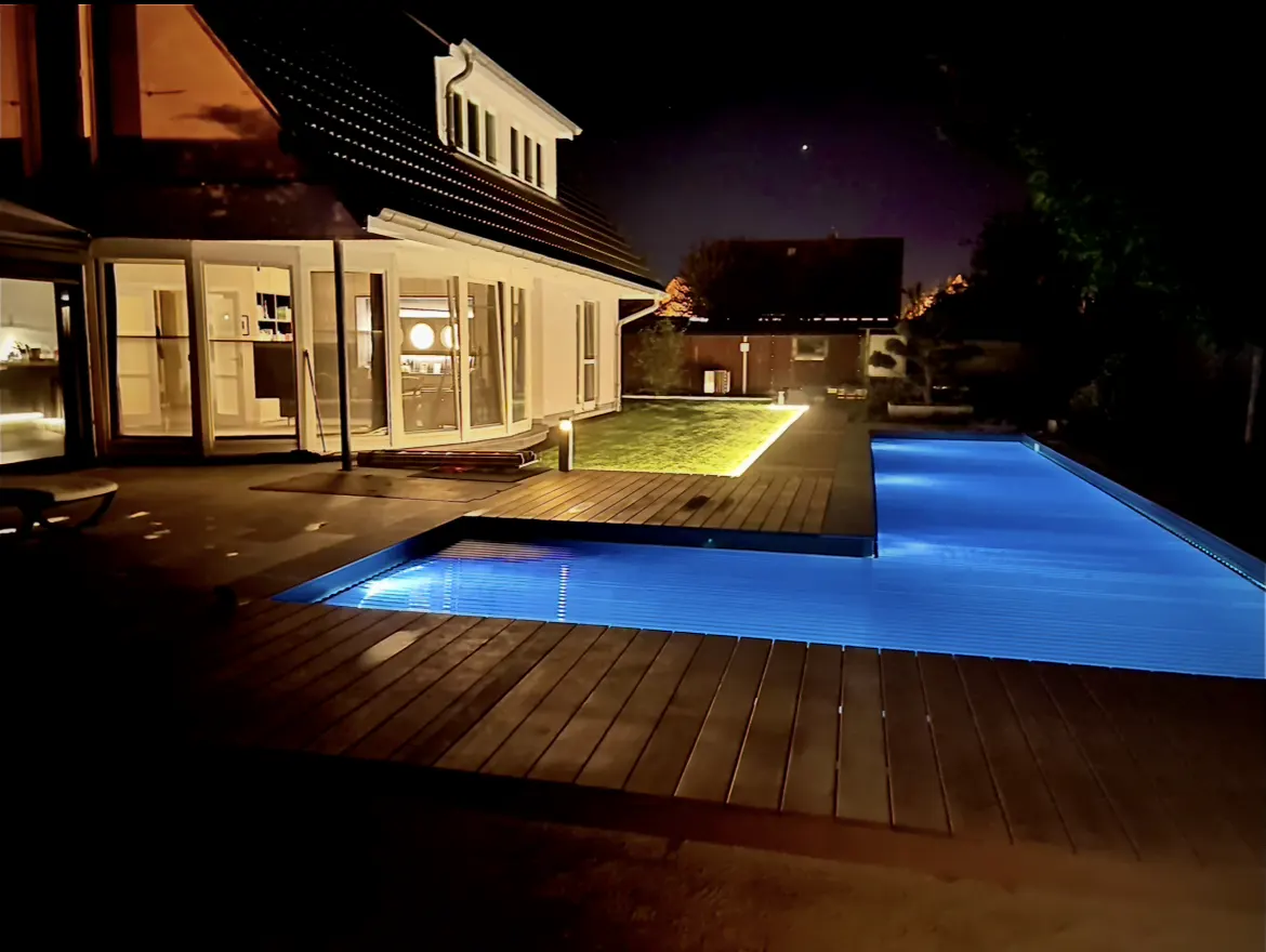 Night view of a house with a pool. The pool is illuminated with blue lights. The house has large glass doors and windows.