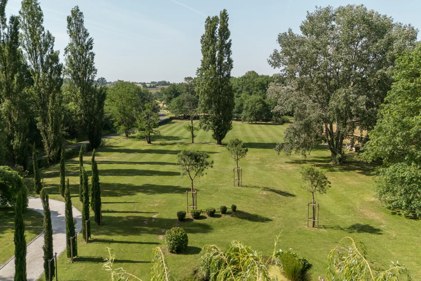 A large green lawn with trees and shrubs, some in protective frames, stretches into the distance under a blue sky.