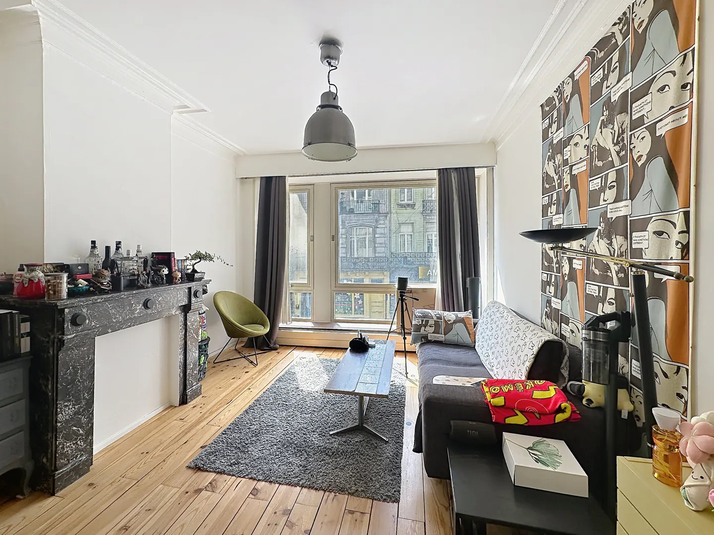 Bright living room with wood floors, a gray sofa, and a comic-style wall hanging. A black fireplace and a green chair add character.