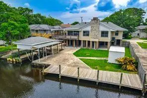 Waterfront home with a dock and boat lift. The beige house has a stone chimney, and is elevated on pilings. Green grass and trees surround the property.