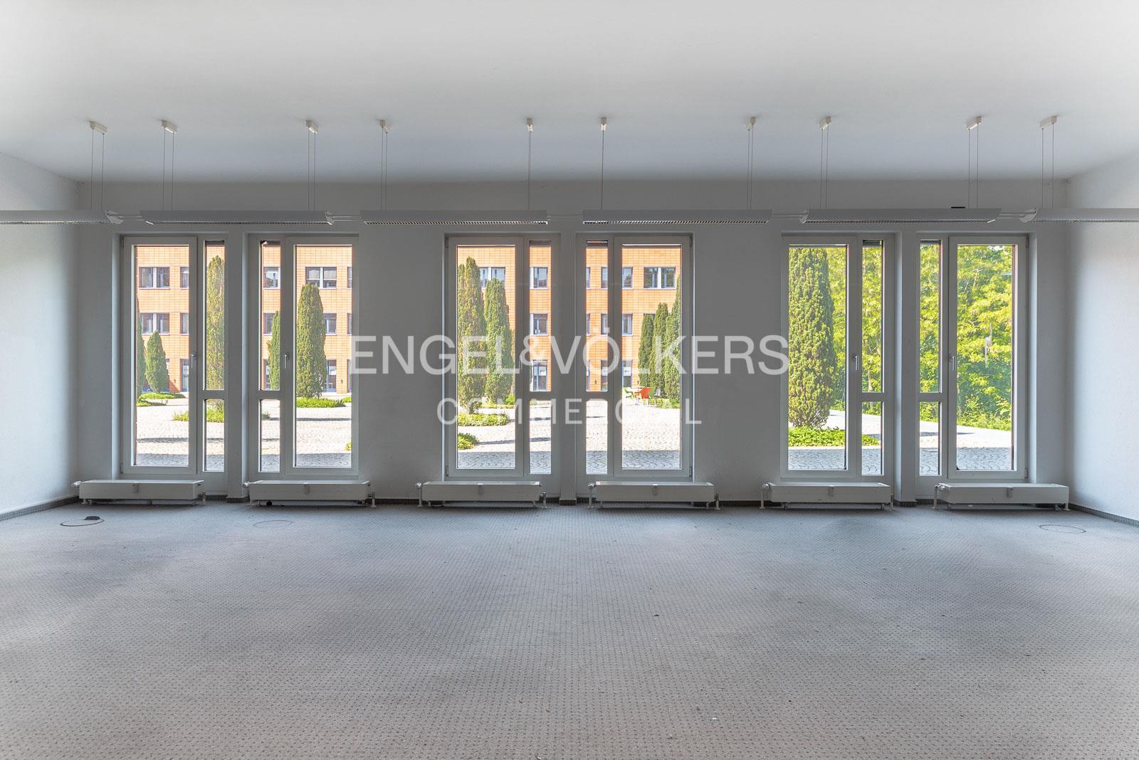 Empty office space with grey carpet, white walls, and large windows overlooking a courtyard with trees and a brick building.