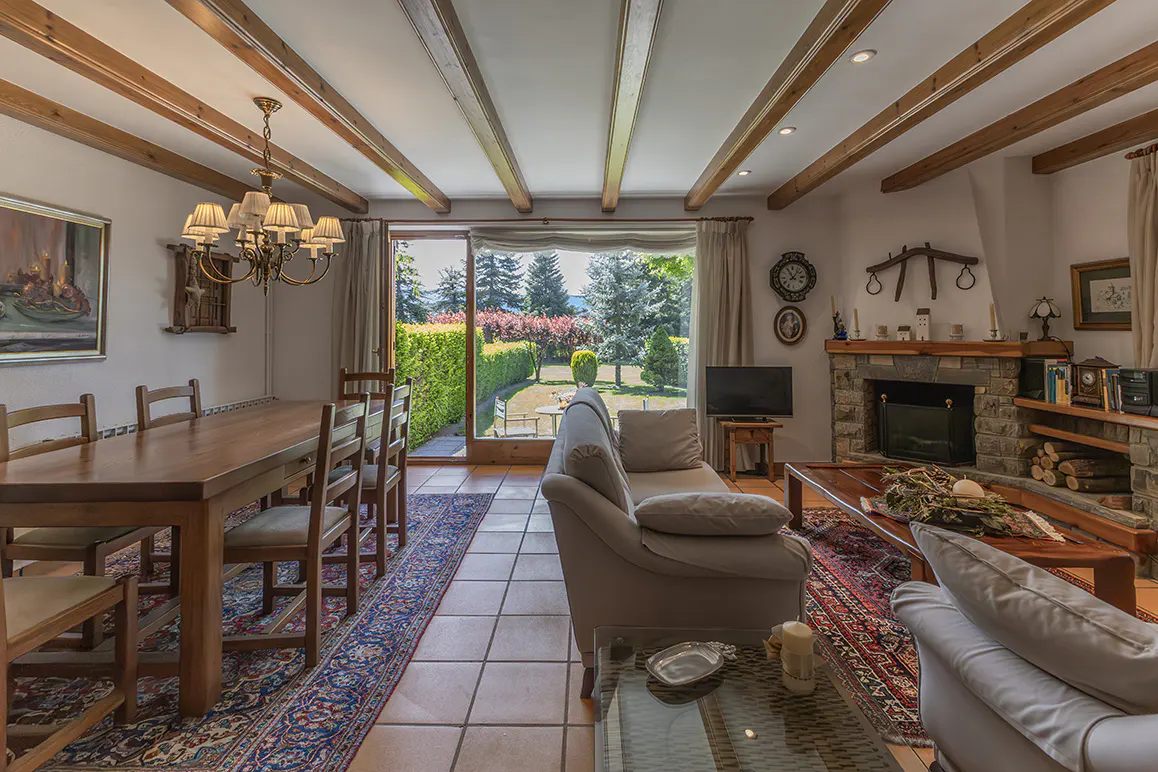 Living room with wooden beams, stone fireplace, sofas, and a dining table. A large window overlooks a green garden.