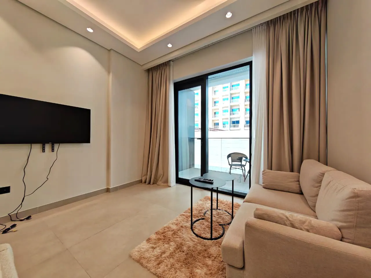 Living room with beige sofa, black TV, and balcony. A round black table sits on a shaggy rug. Curtains frame the sliding glass door to the balcony.