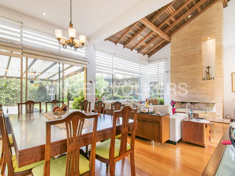 Bright, open dining room with a large wooden table, chairs, and chandelier. Floor-to-ceiling windows overlook a lush garden. A brick fireplace adds warmth.