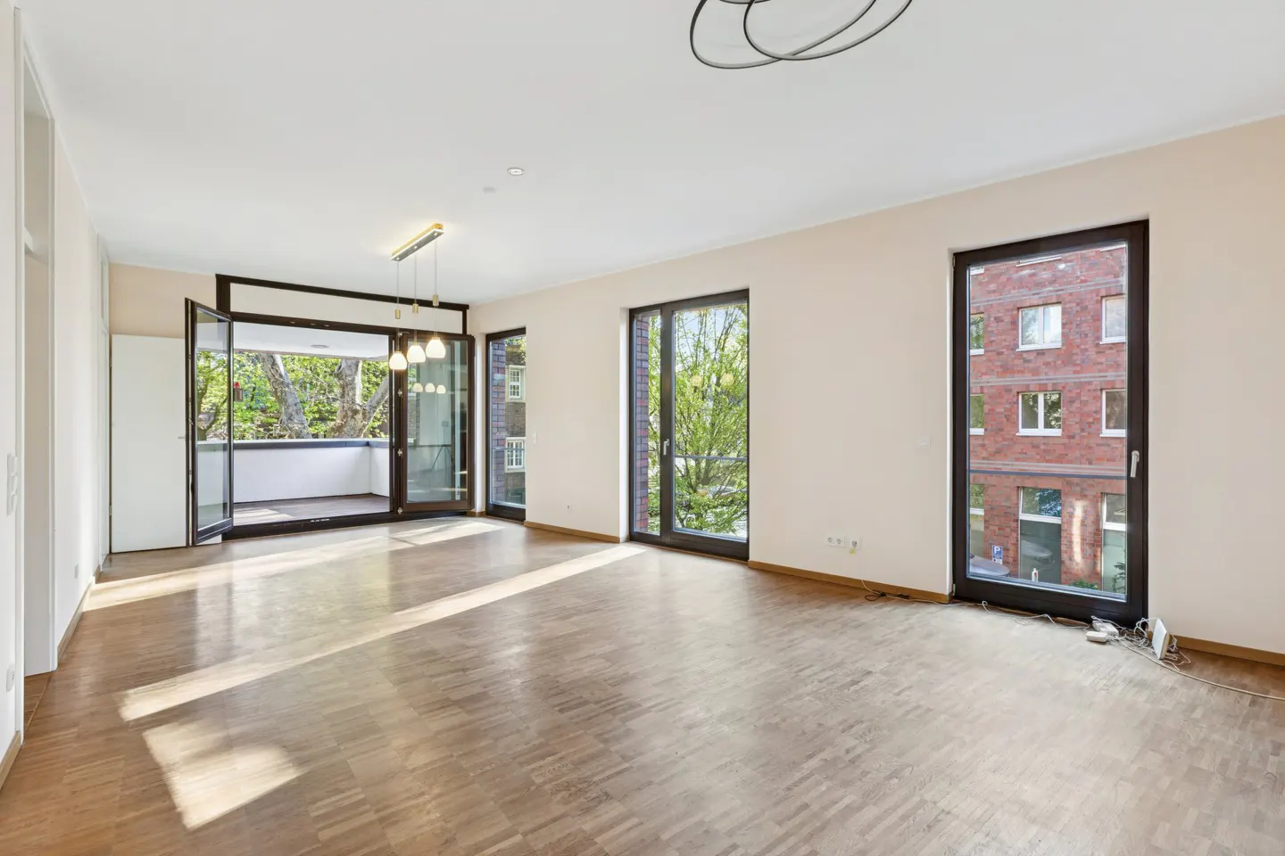Bright, empty living room with wood floors, white walls, and black-framed windows. Balcony doors open to trees. Modern light fixture on ceiling.