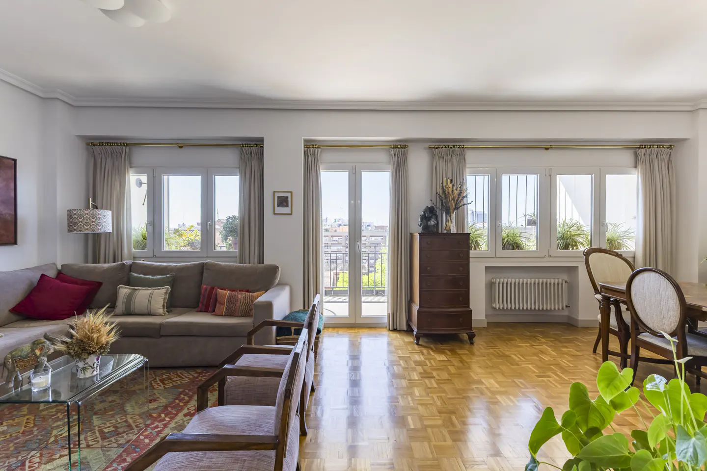 Bright living room with a brown sectional sofa, a glass coffee table, and a wooden floor. Windows with curtains overlook a city view.