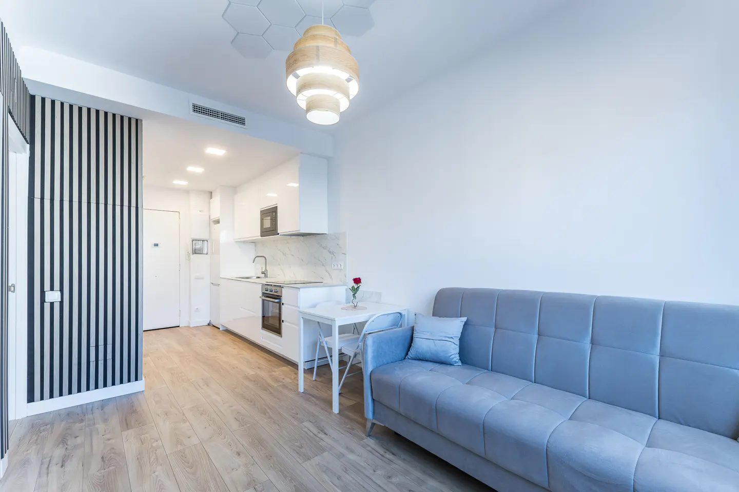 Bright, modern apartment with a blue sofa, white kitchen, and wood floors. A wooden chandelier hangs above a small white table with a single red rose.