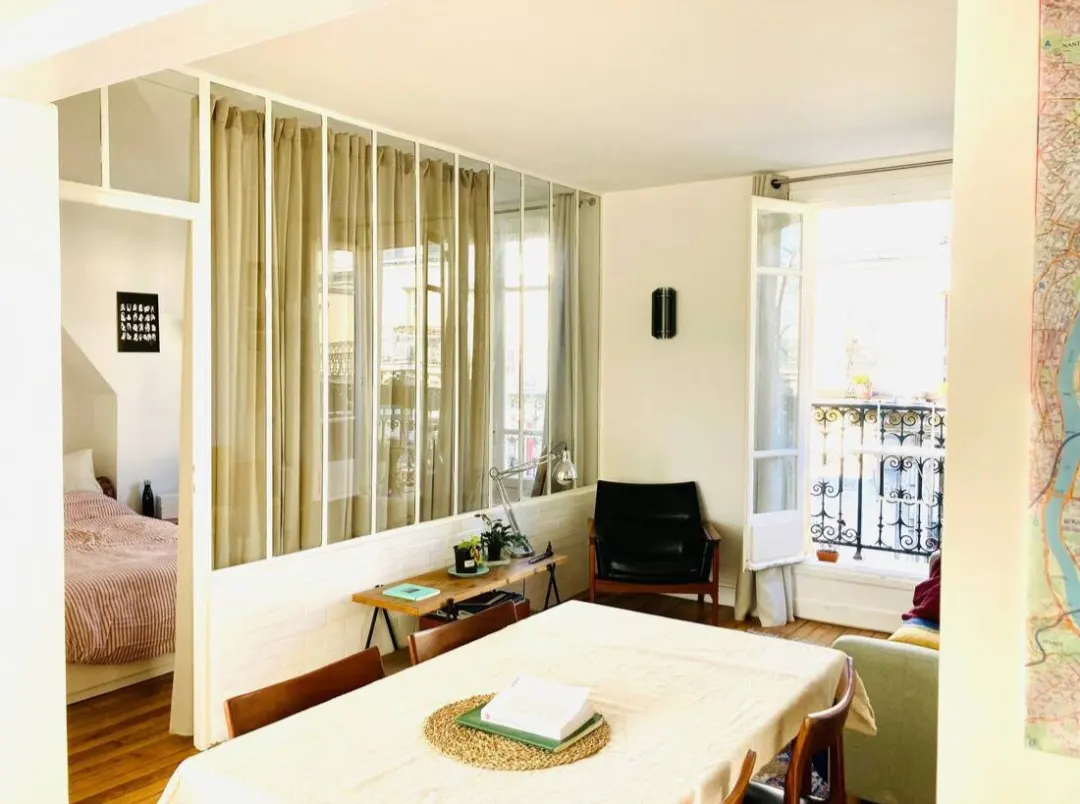 Bright apartment interior with a dining table, chairs, and a glass-paneled wall with curtains separating the bedroom. Balcony visible.