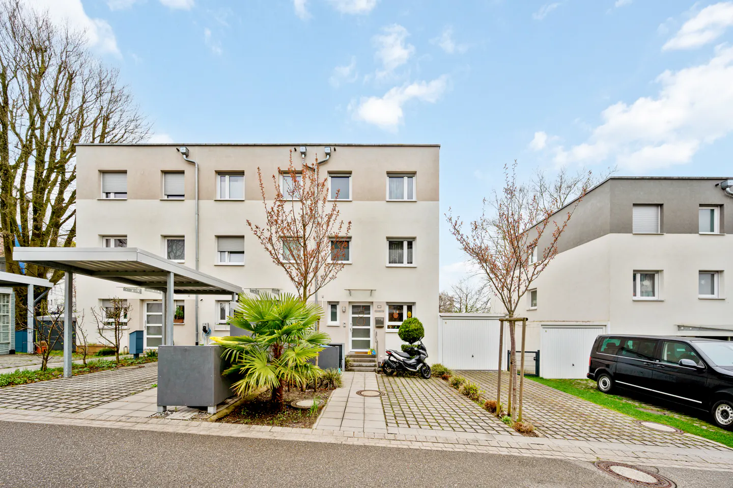 Exterior view of a modern, three-story apartment building with a scooter parked in front. A black van is parked on the right.