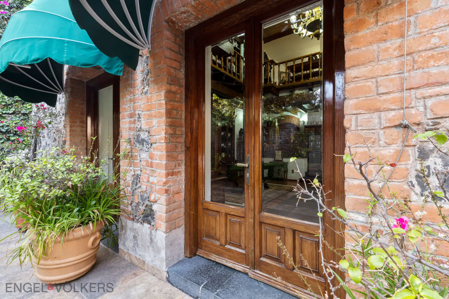Exterior view of a brick building with a wooden door and a green awning. A potted plant sits near the entrance.