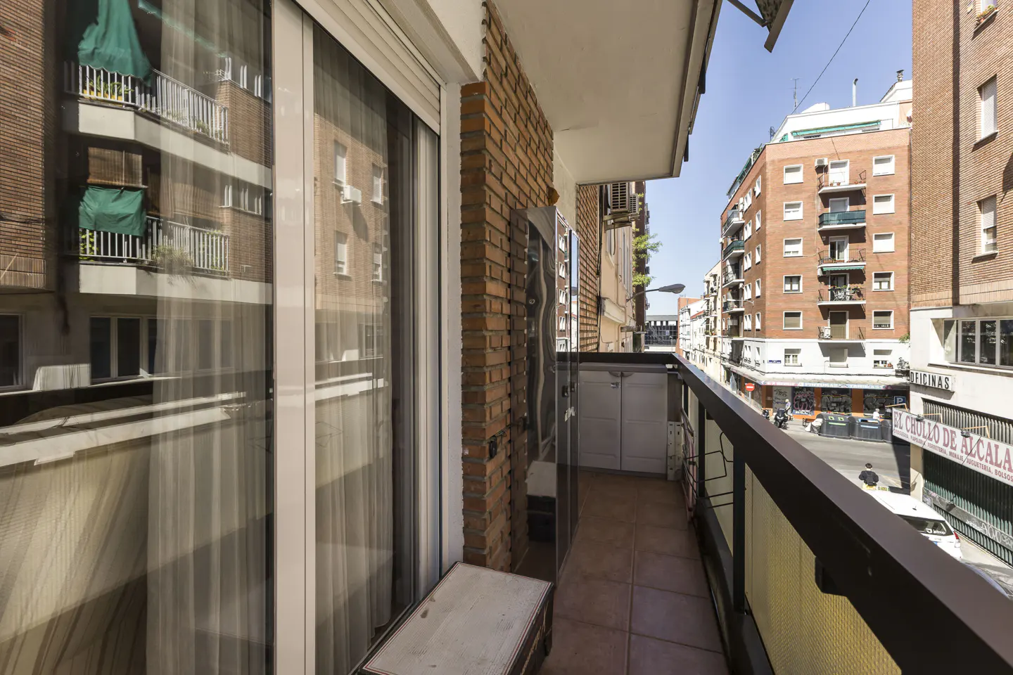 A narrow apartment balcony with a brick wall, tiled floor, and metal railing overlooking a sunny city street.