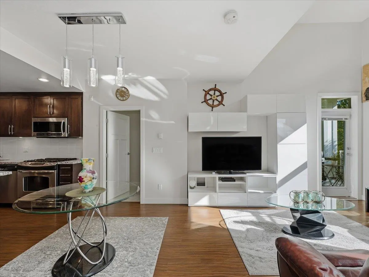Open-concept living room with wood floors, glass dining table, white media console, TV, and kitchen with dark cabinets.