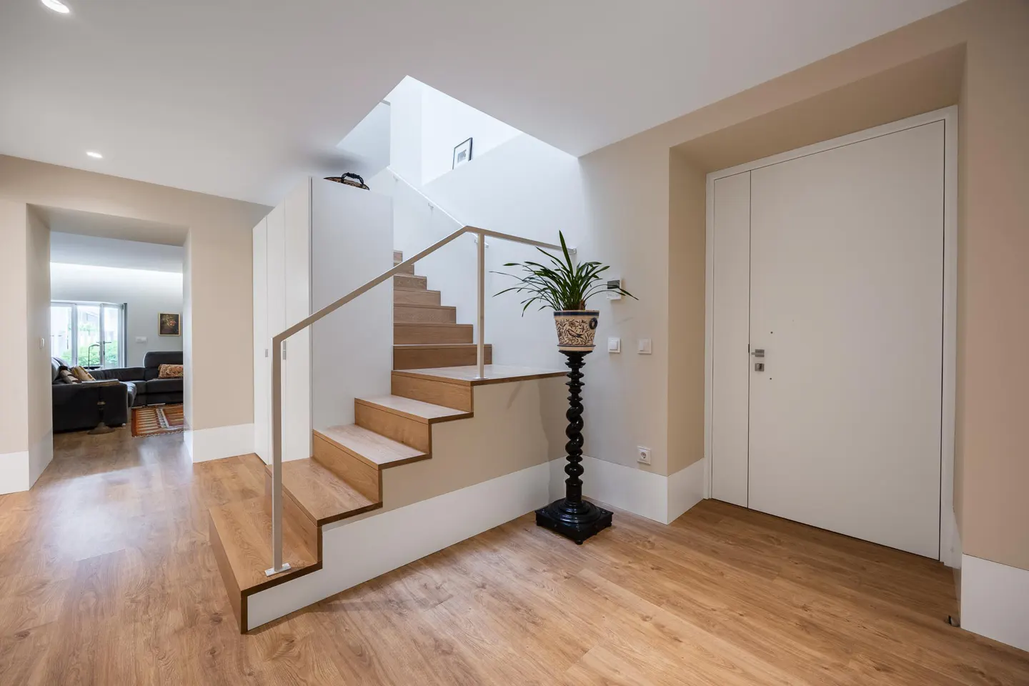 Interior view of a home's entryway with wood floors, stairs with metal railing, and a white door. A plant sits on a black stand.