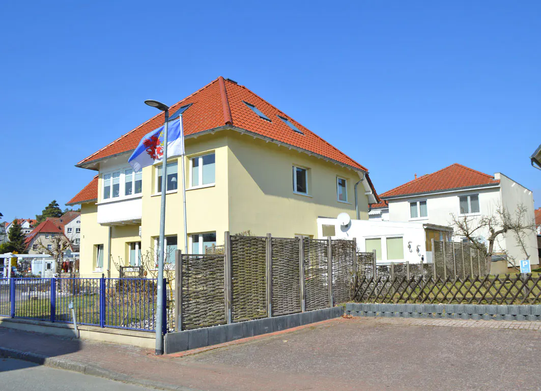 Two-story yellow house with a red tile roof and white trim, behind a woven fence and blue metal fence. Flags are flying in front.