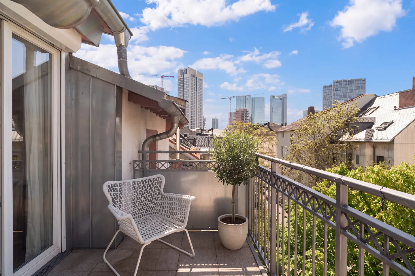 Balcony view with white wicker chair, potted tree, and metal railing overlooking city buildings under a blue sky.