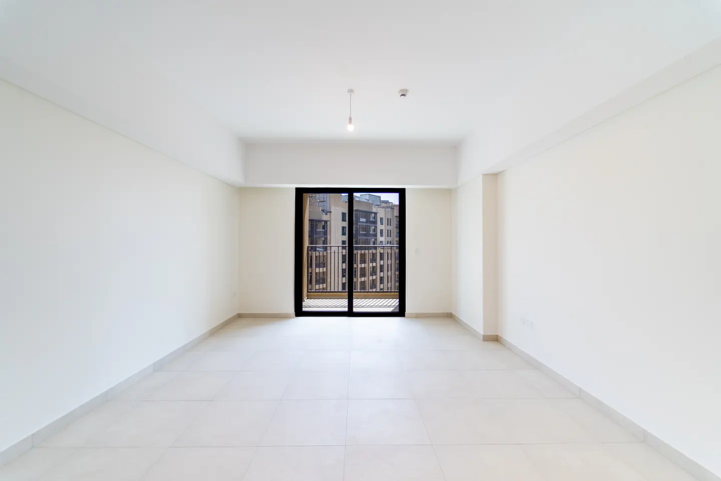 Bright, empty room with white walls and tile floor. Black-framed sliding glass doors lead to a balcony with a city view.
