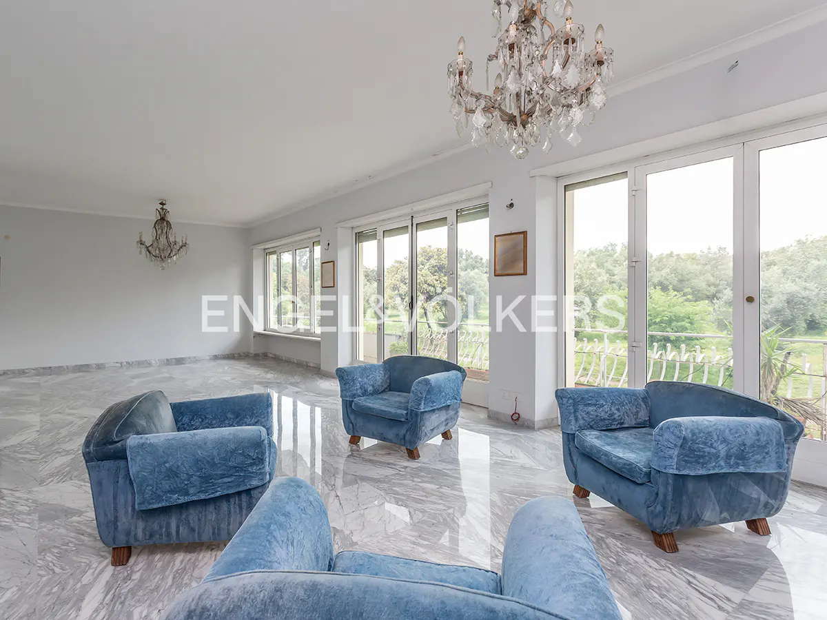 A bright living room with blue armchairs, marble floors, and a chandelier. Large windows overlook a green landscape.