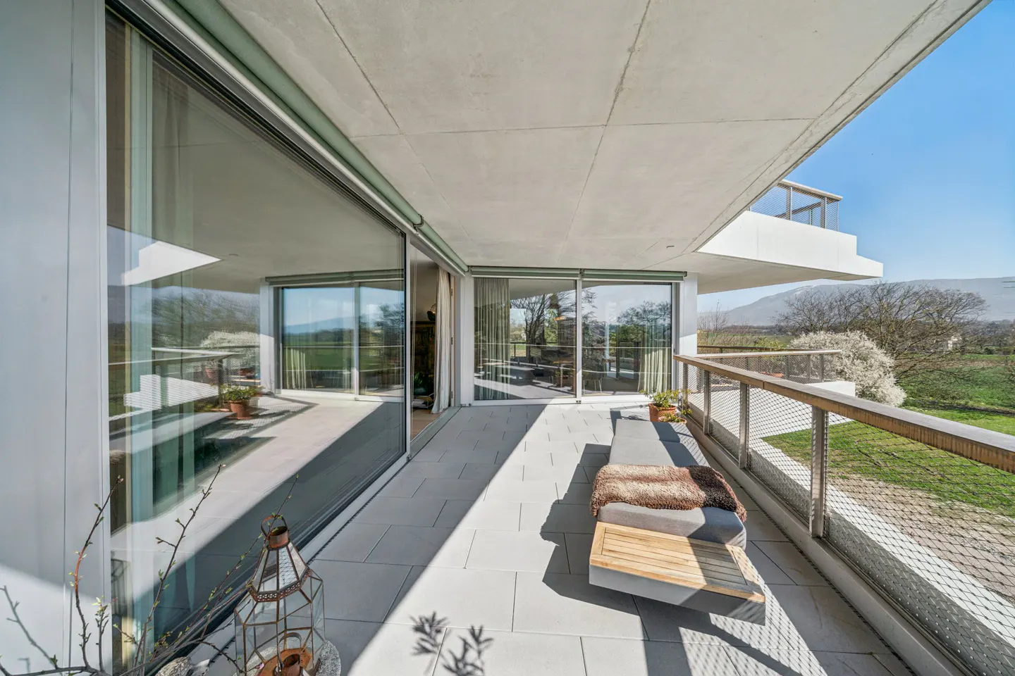 Modern balcony with gray tile floor, glass doors, and wood railing overlooking green fields and trees under a blue sky.