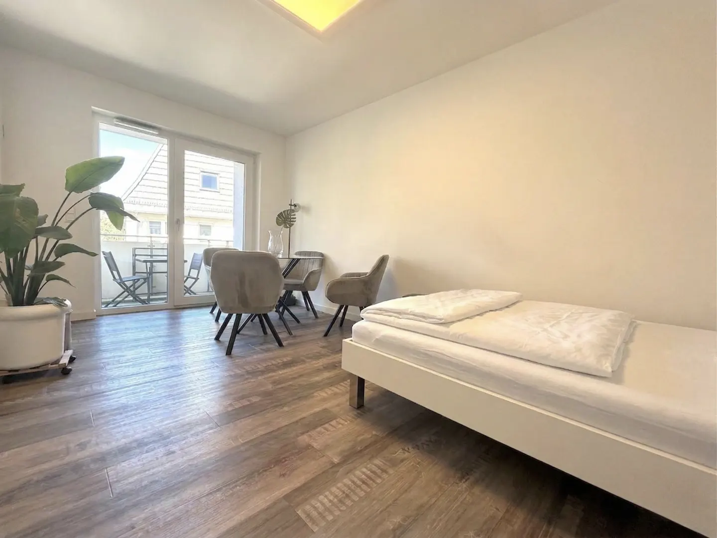 A bright, modern apartment with wood floors, a white bed, a dining table with four chairs, and a balcony with outdoor furniture.
