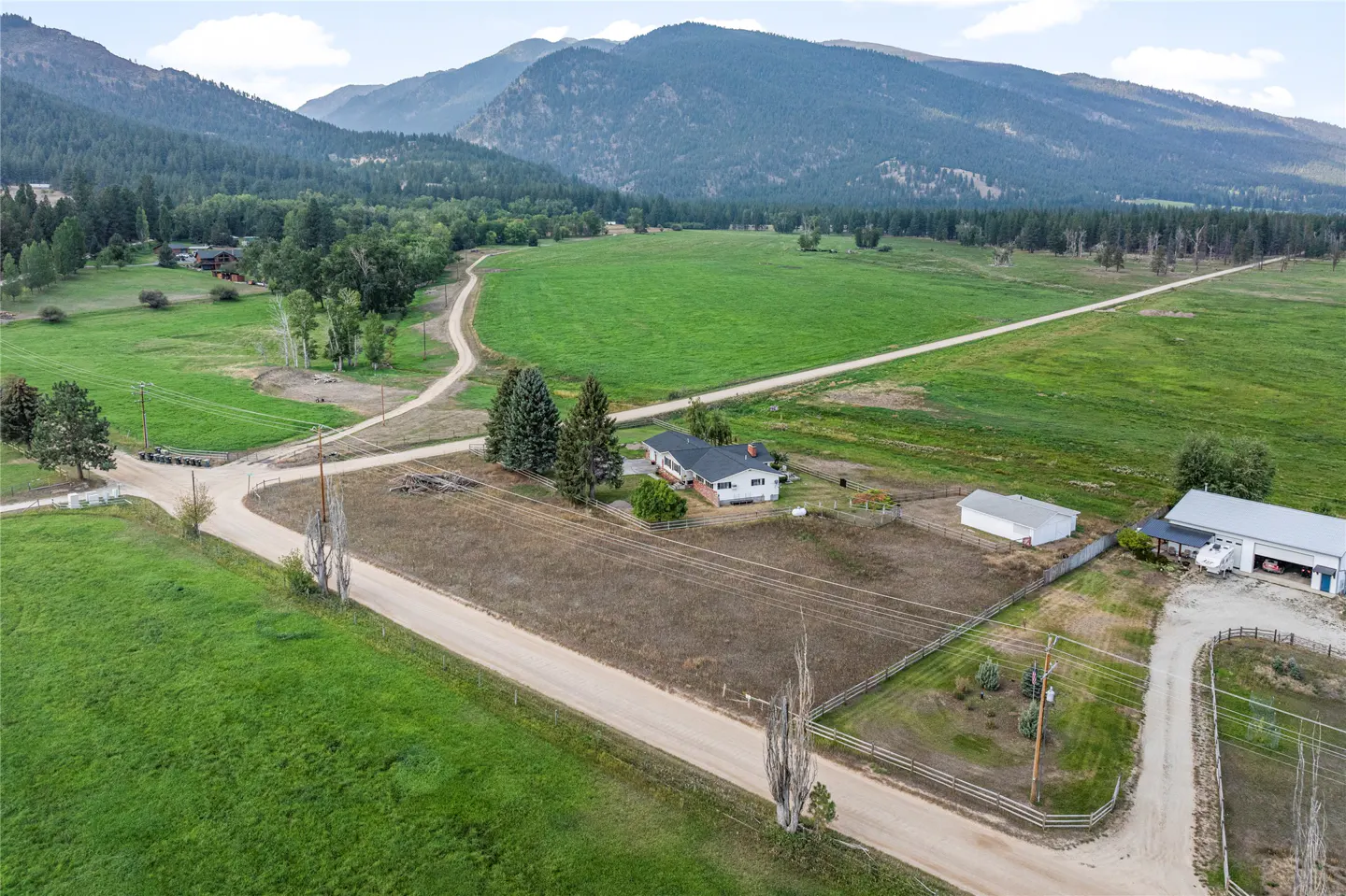 Aerial view of a white house with outbuildings on a large lot with green fields and mountains in the background.