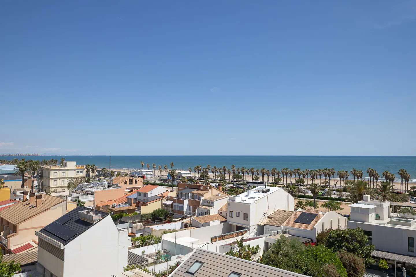 A sunny, high-angle view of a coastal town with white buildings, palm trees, and the blue ocean under a clear sky.