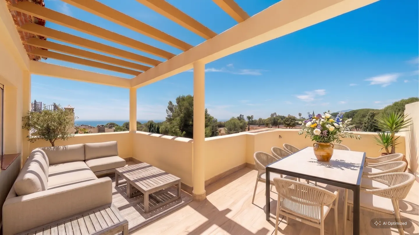 Outdoor patio with beige furniture, a wooden pergola, and a view of trees and the ocean under a blue sky. A table with flowers sits to the right.