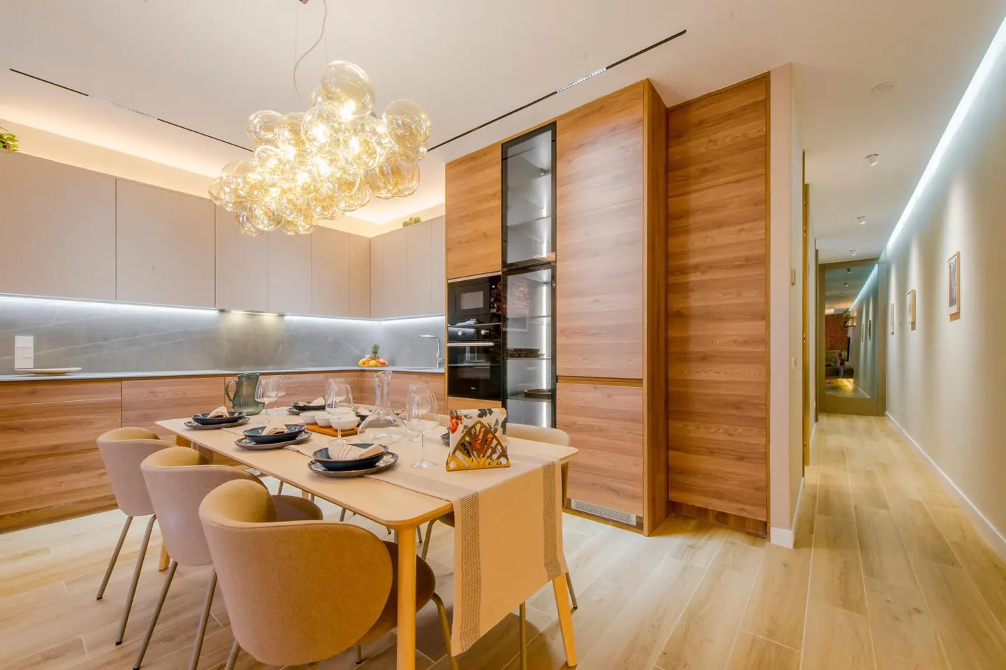 A modern kitchen and dining area with wood cabinets, a glass chandelier, and a long dining table with six chairs.