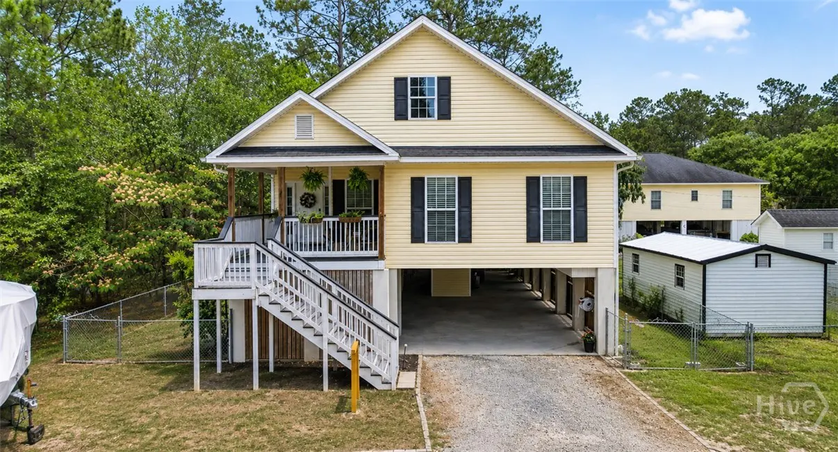 Two-story yellow house with black shutters, elevated on stilts. White stairs lead to a porch with plants. Trees and blue sky in the background.