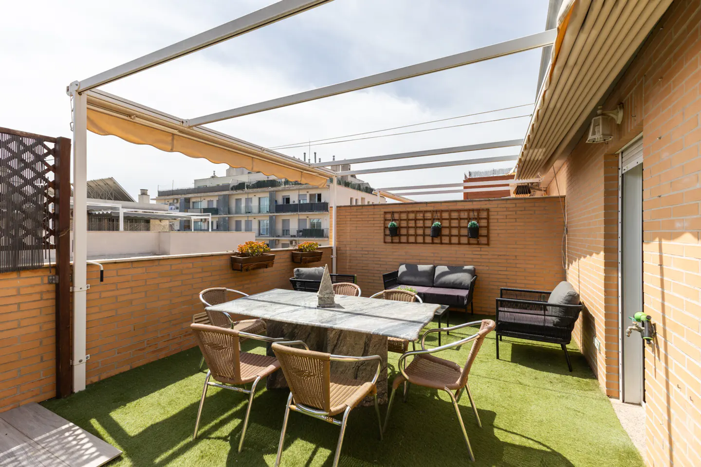Rooftop patio with green turf, brick walls, and a white pergola. A stone table with wicker chairs sits near a gray sofa and black armchair.