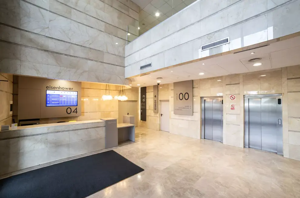 Modern office lobby with marble walls and floors, a reception desk with a screen, and two elevators.