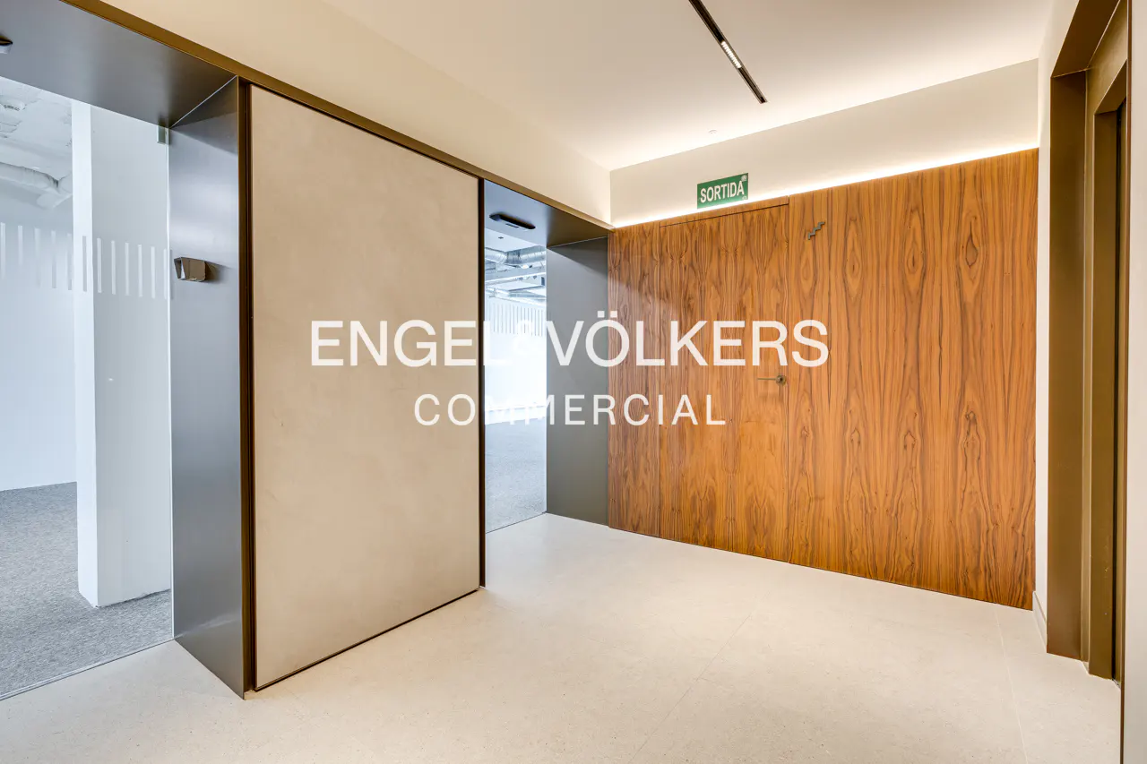 Office interior with Engel & Völkers Commercial logo on a beige wall. Wood-paneled wall with "Sortida" sign. White tile floor.