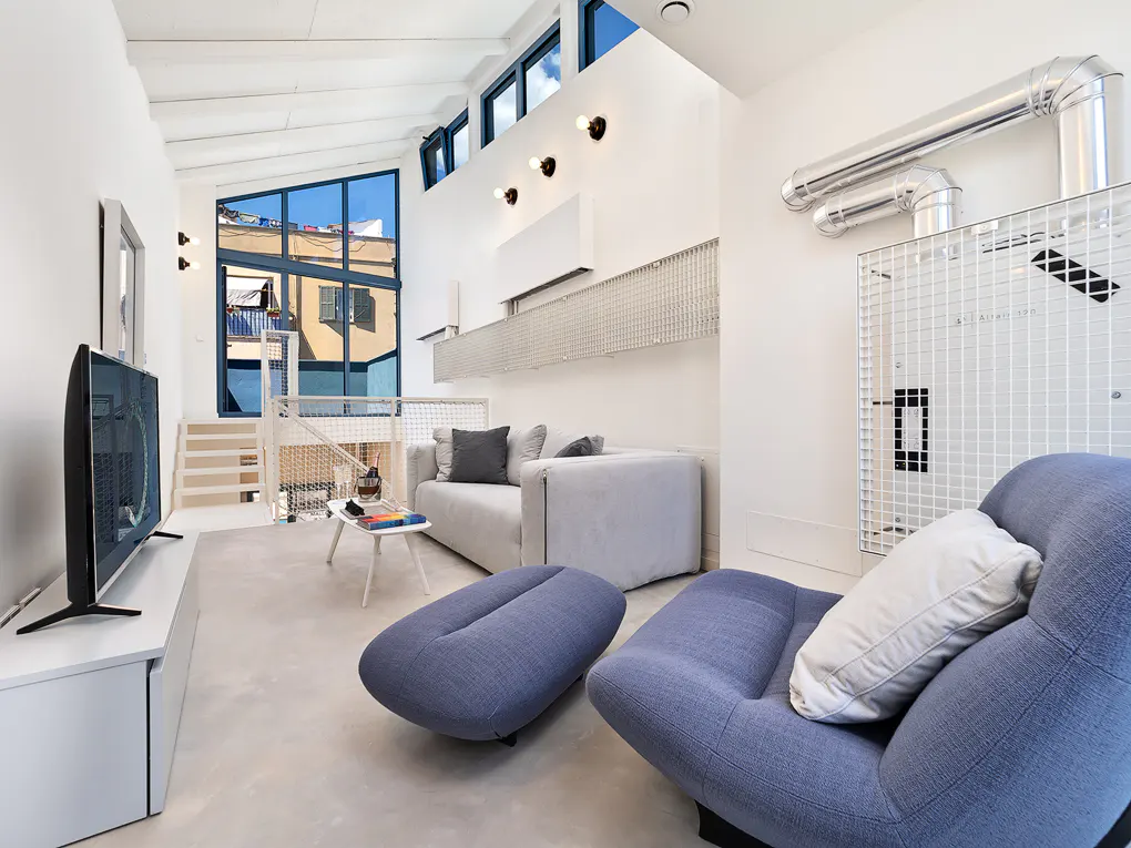 Bright, modern living room with white walls, blue chair and ottoman, grey sofa, and TV. Large window shows a building exterior.