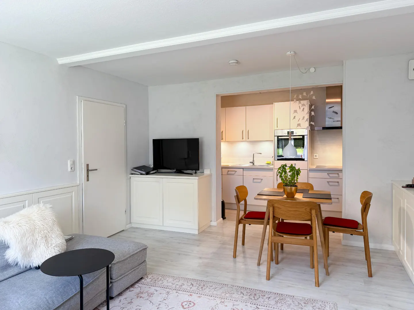 Open-concept living space with a gray sofa, black table, and a dining table with four chairs. The kitchen has white cabinets.