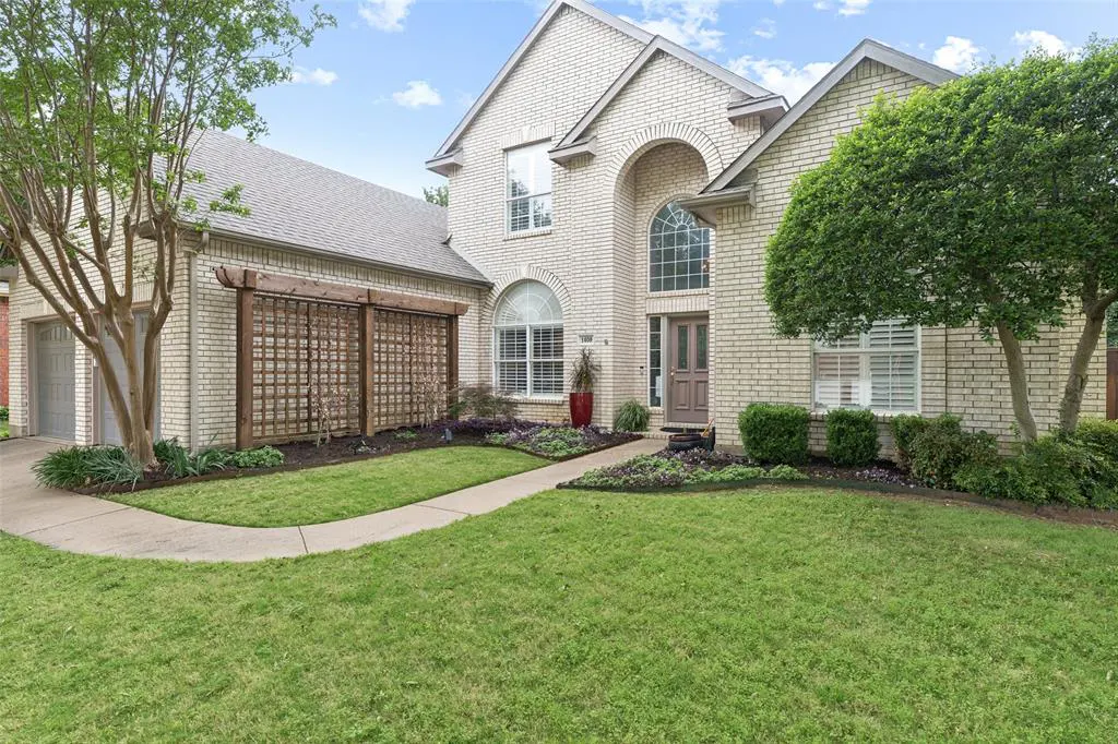 Two-story beige brick home with a manicured lawn, walkway, and a wooden trellis on the left.